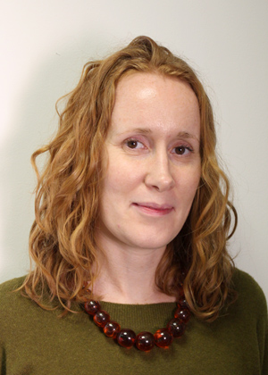 Headshot photo of Suzi Mackenzie. White female with auburn curly long hair and brown eyes. Wearing an amber necklace and green jumper