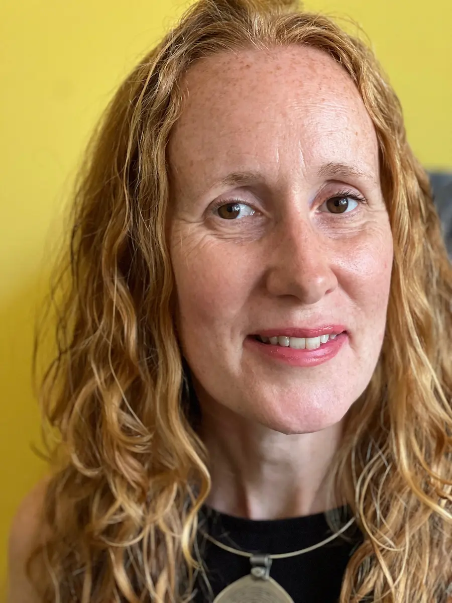 A headshot photograph of Suzi smiling at the camera. She has curly red long hair and brown eyes. She is wearing a black top and large silver necklace.
